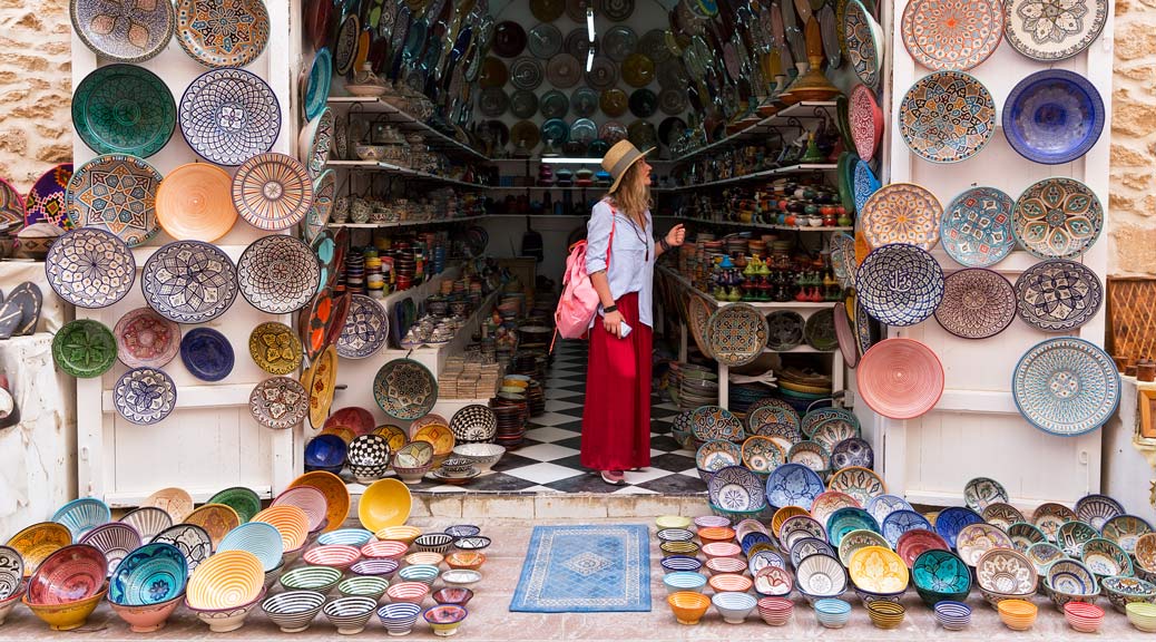 Young blonde woman in a long red skirt, hat and with pink backpack is standing in a small shop and looking at Moroccan colorful plates.