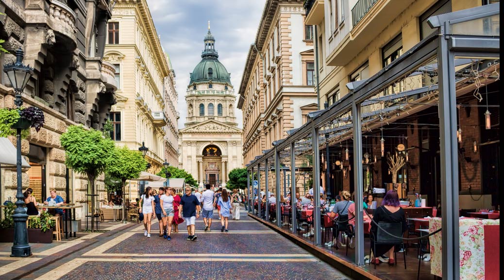 tourists take a stroll next on a cobbled street in Budapest next to a outdoor cafe