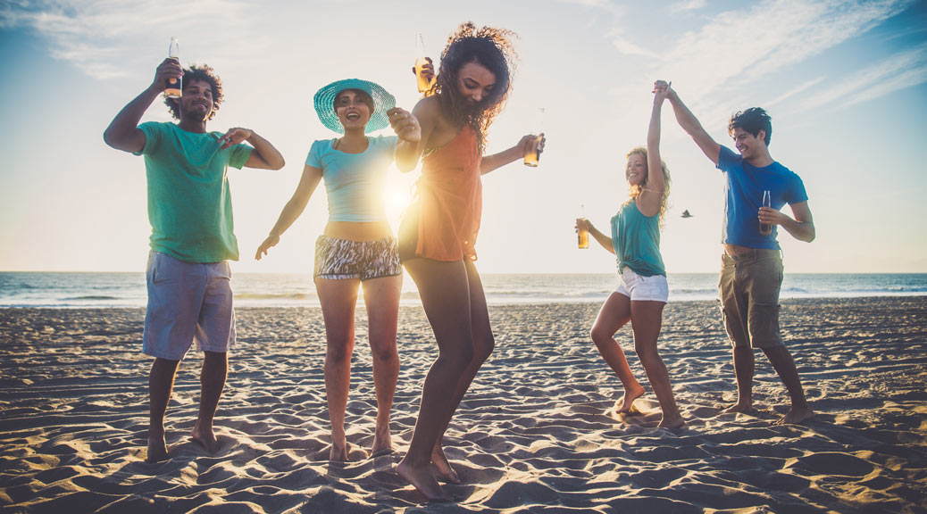 group of teenagers enjoying and dancing holding beer bottles in hand during sunset at the beach