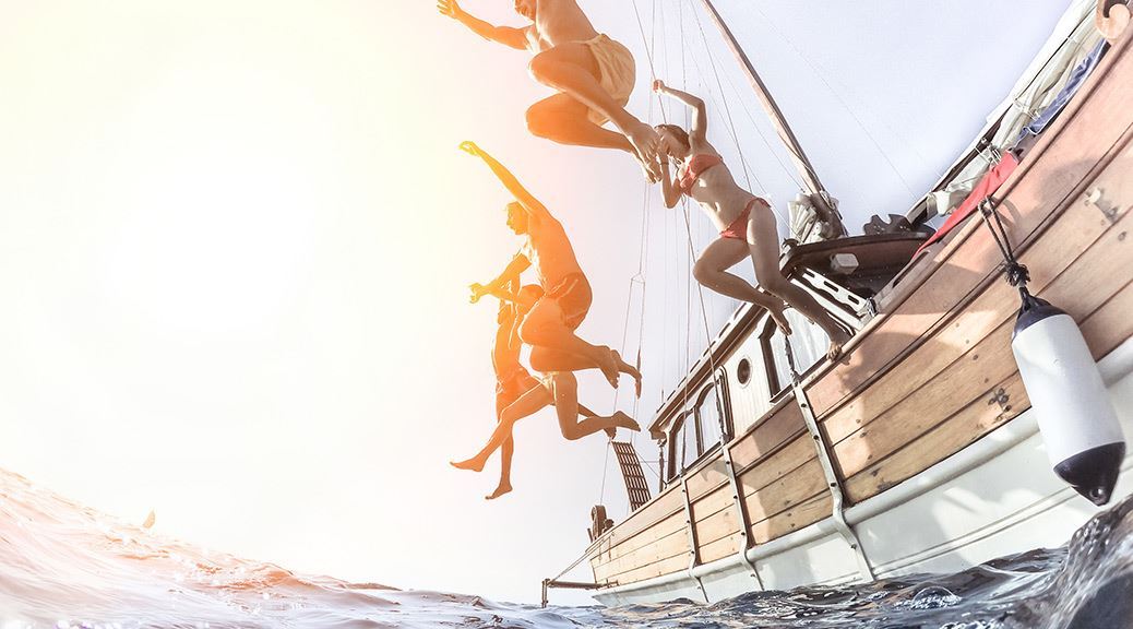 group of teenagers jumping out of the sail boat into the sea during a sunset